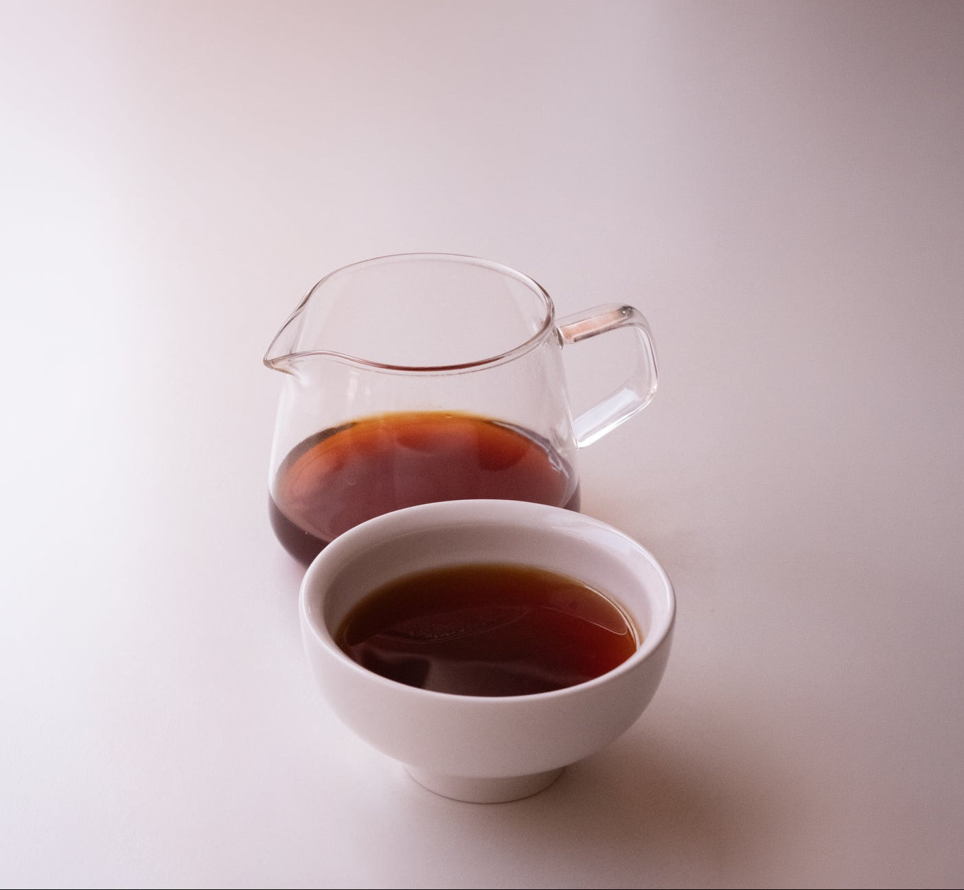 A small clear glass pitcher and a white ceramic cup filled with dark amber-brown coffee, photographed on a clean off-white background.